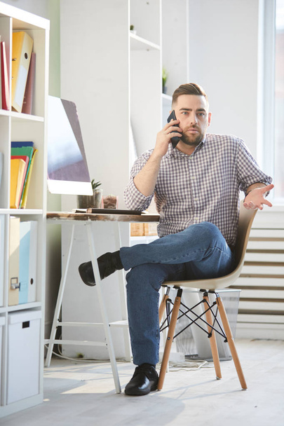 junger zeitgenössischer Geschäftsmann in Freizeitkleidung sitzt auf Stuhl an Schreibtisch und berät sich am Telefon im Büro - Foto, Bild