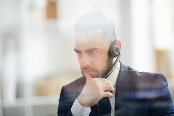 Homme d'affaires sérieux dans casque écoutant attentivement l'un des clients en ligne tout en étant assis par le lieu de travail
 - Photo, image