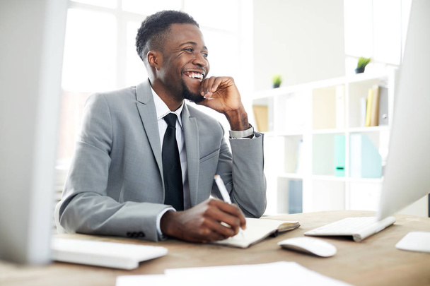Szczęśliwy młody African-American Manager w formalwear śmiech na coś śmiesznego na ekranie komputera siedząc w miejscu pracy - Zdjęcie, obraz