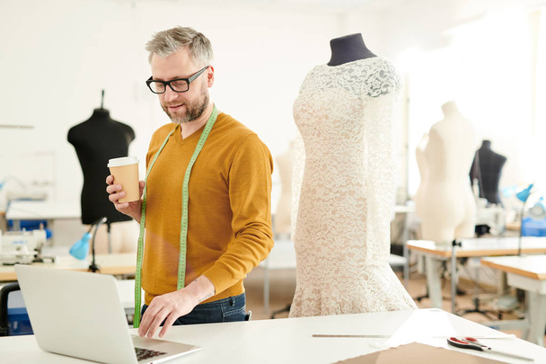 Dojrzały profesjonalny projektant mody z taśmą pomiarową i kieliszek napoju stojącego przy biurku przed laptopem w studiu - Zdjęcie, obraz