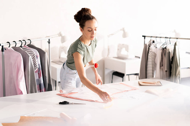 Mujer atractiva joven en ropa de casualwear de pie por la mesa mientras toma pines fuera de patrón de papel fijado hasta textil
 - Foto, Imagen