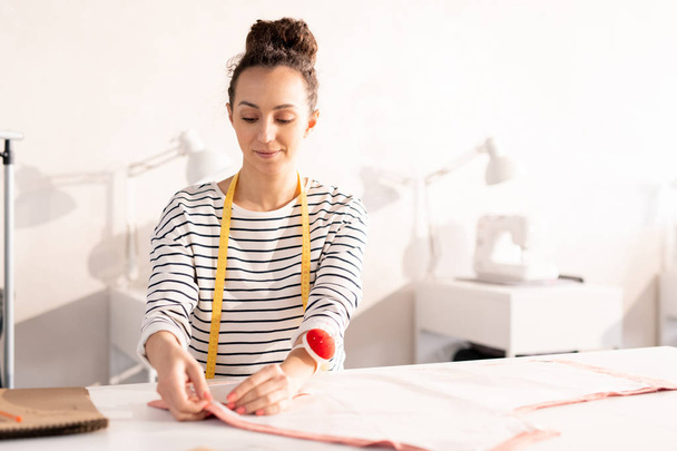Young seamstress or fashion designer in casualwear pinning paper pattern on piece of fabric during work over new order of client - Foto, afbeelding