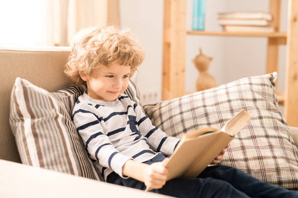 Cute and clever young boy in casualwear sitting in comfortable armchair and reading book of tales - Foto, Imagem