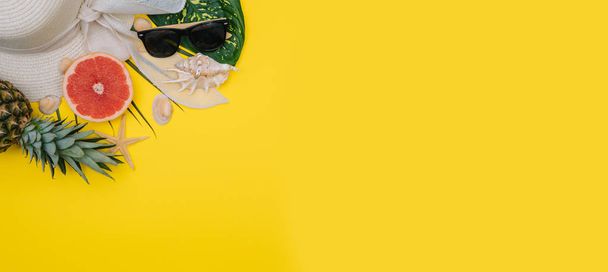 Flat lay beach accessories: swimsuit, sunglasses, hat, sandals with palm branches, exotic fruits and shells. Top view on yellow background - 写真・画像