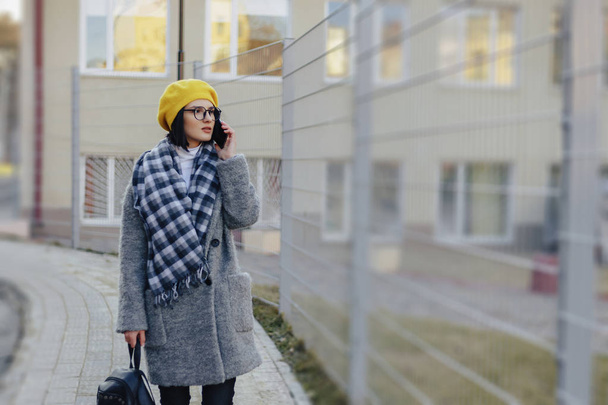 Привлекательная молодая девушка в солнечных очках в пальто, идущая по улице, разговаривающая по телефону и улыбающаяся
 - Фото, изображение