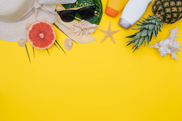 Beach flat lay accessories. Sun hat, towel, camera, cream, swimsuit, slippers, sunscreen bottle, exotic fruits, and seashells on a yellow background. Summer travel holiday concept - 写真・画像