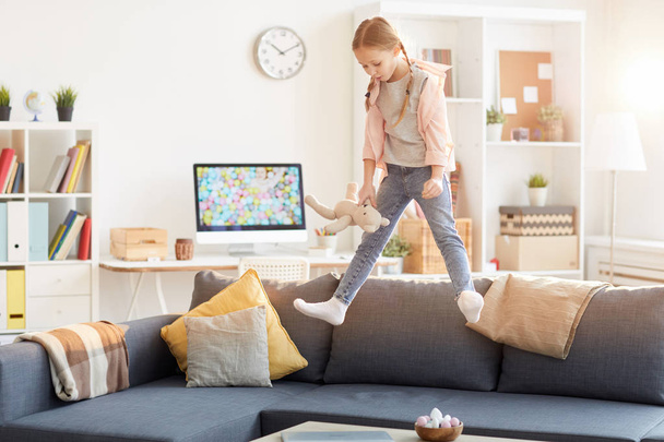 Full length portrait of cute girl with pigtails jumping on couch at home lit by sunlight, copy space - Photo, Image