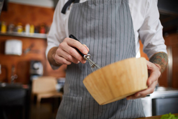 Střední část portrétu neidentifikovatelného kuchaře mixování něčeho v dřevěné misce se šlehací metou - Fotografie, Obrázek