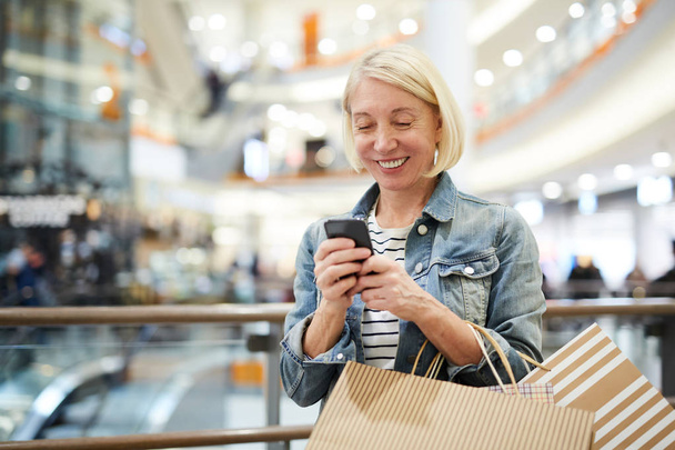 Feliz mulher madura animado com cabelo loiro curto em pé no corredor do shopping center e SMS no smartphone enquanto conversa on-line com assistente de compras
 - Foto, Imagem