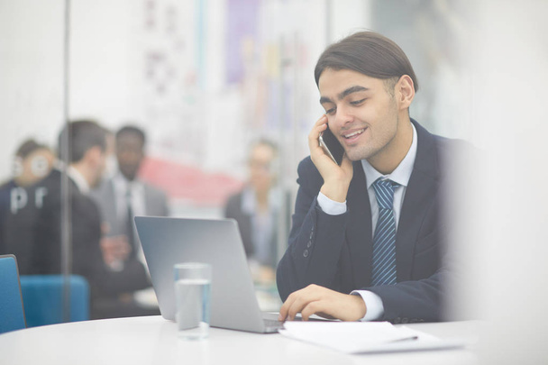 Портрет молодого ближневосточного бизнесмена, говорящего по телефону и пользующегося ноутбуком во время работы в офисе, снятого из-за стекла, копировального пространства
 - Фото, изображение