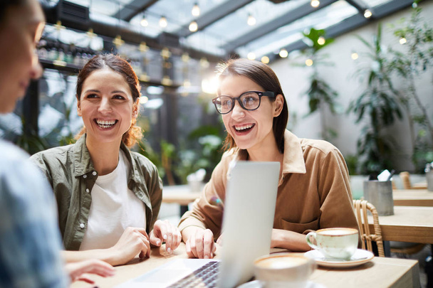 Позитивні схвильовані молоді жінки-колеги в повсякденному одязі сидять за столом в сучасному кафе і сміються під час роботи в якості команди в кафе
 - Фото, зображення
