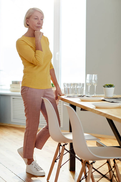 Σοβαρή γυναικεία ξανθιά ώριμη γυναίκα σε casual ρούχα στέκεται στο τραπέζι του φαγητού και γέρνει το κεφάλι στο χέρι, ενώ σκέφτονται για τη ρύθμιση του τραπεζιού στο σπίτι - Φωτογραφία, εικόνα