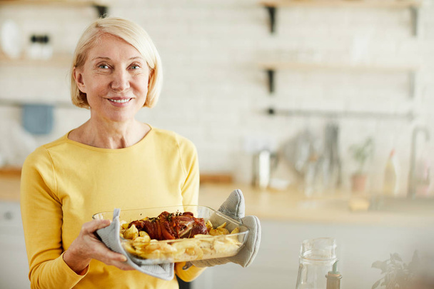 fröhlich attraktive vorsichtige ältere Dame mit kurzen blonden Haaren steht in der modernen Küche und hält Schüssel mit gekochtem Fleisch, während sie in die Kamera schaut - Foto, Bild