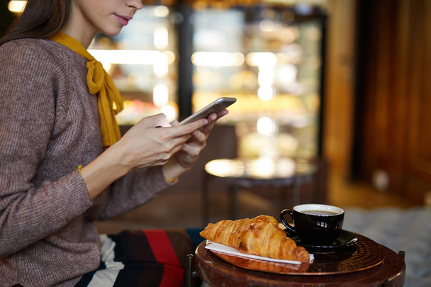 Spokojny młoda kobieta ze smartfonem szuka kontaktu siedząc przy stole w kawiarni i mając ciasto z herbatą - Zdjęcie, obraz