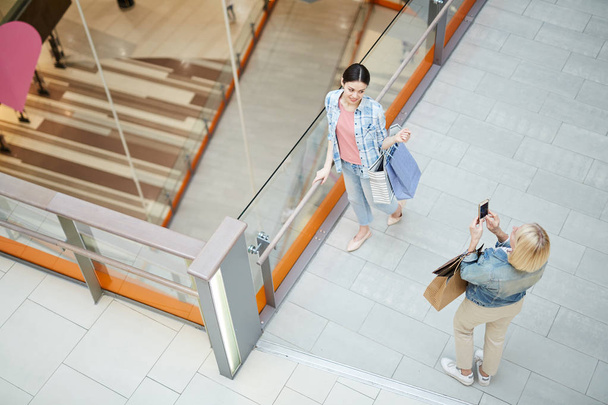 Top view of blond lady using smartphone to photograph friend with shopping bags in modern mall - Photo, Image