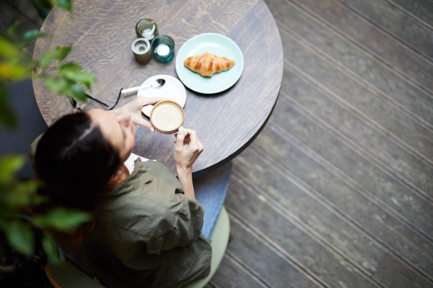 コーヒーショップで朝食をとりながらテーブルに座り、カプチーノを飲む若い女性の眺めの真上 - 写真・画像