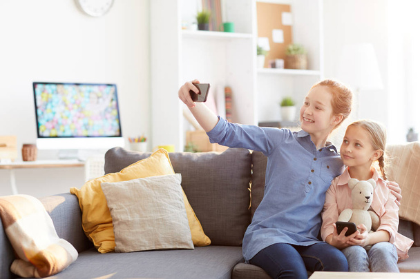 Portret dwóch sióstr robienia selfie razem siedząc na kanapie w domu i uśmiechając się do kamery mobilnej, Kopiuj przestrzeń - Zdjęcie, obraz