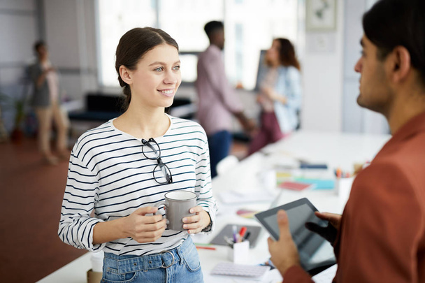 Taille up portret van hedendaagse jonge vrouw praten met collega en glimlachend tijdens koffiepauze in kantoor, kopieer ruimte - Foto, afbeelding