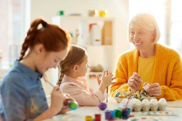 Portrait of smiling mature woman talking to little girl while painting Easter eggs together in art class, copy space - Photo, Image