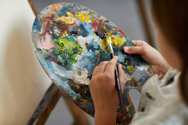 Close-up van onherkenbaar meisje Holding artiesten palet en mengen van kleuren tijdens het schilderen op ezel in kunst klasse, kopieer ruimte - Foto, afbeelding