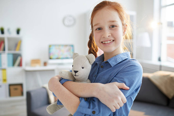 Waist up portrait of red haired teenage girl hugging plush toy and smiling at camera, copy space - Photo, image