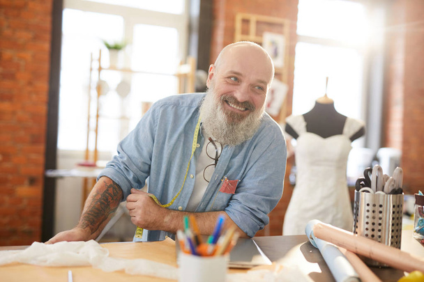 Boldog szakállas Tailor a casualwear állva az asztalra, miközben beszél kollégája a stúdióban a divattervezés - Fotó, kép