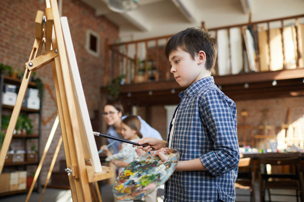 Ritratto vista laterale di smart schoolboy pittura quadro sul cavalletto in classe d'arte e tenendo tavolozza, spazio copia
 - Foto, immagini