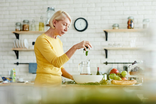 Ernstige aantrekkelijke rijpe vrouw in gele trui staande op de teller en het toevoegen van spinazie in kom, terwijl het maken van gezonde salade in de binnenlandse keuken - Foto, afbeelding
