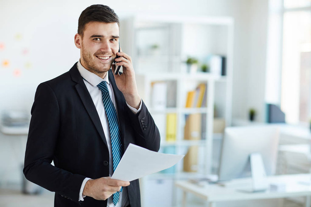 Щасливий молодий успішний банкір у формальному одязі розмовляє з одним з клієнтів по телефону в офісі
 - Фото, зображення