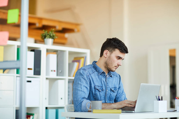 Vakava nuori toimistotyöntekijä satunnaisissa vaatteissa istuu pöydän ääressä kannettavan tietokoneen edessä ja työskentelee online-tietojen kanssa
 - Valokuva, kuva