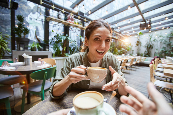 Jó idő a kávézóban: vidám aatractive fiatal nő ül az asztalnál, és intés a kamera csevegés közben a barát és iszik kávét - Fotó, kép
