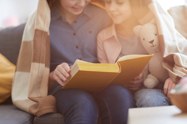 Closeup portrait of two sisters reading book under blanket lit by warm sunlight, copy space - Photo, image