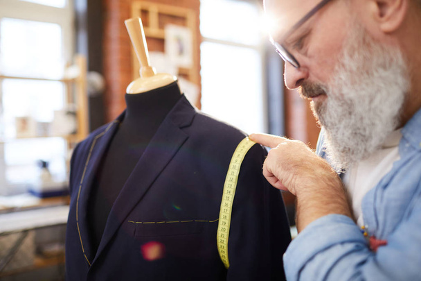 Выдержанный бородатый профессиональный портной с помощью измерительной ленты во время работы над новой курткой в своей мастерской
 - Фото, изображение