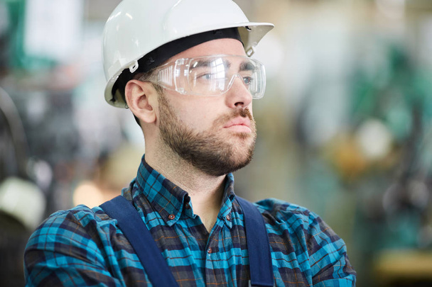 Cabeça e ombros portrit de trabalhador da fábrica barbudo vestindo hardhat olhando para longe pensivamente, espaço de cópia
 - Foto, Imagem