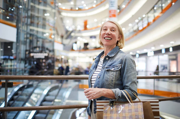 Весела збуджена зріла жінка з світлим волоссям, що стоїть в торговому центрі і сміється, дивлячись на камеру
 - Фото, зображення