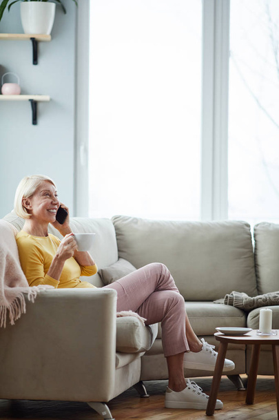 Χαρούμενα ώριμη ξανθιά γυναίκα σε casual ρούχα κάθεται σε άνετο καναπέ στο σαλόνι και μιλάει στο τηλέφωνο, ενώ πίνοντας καφέ από το φλιτζάνι - Φωτογραφία, εικόνα
