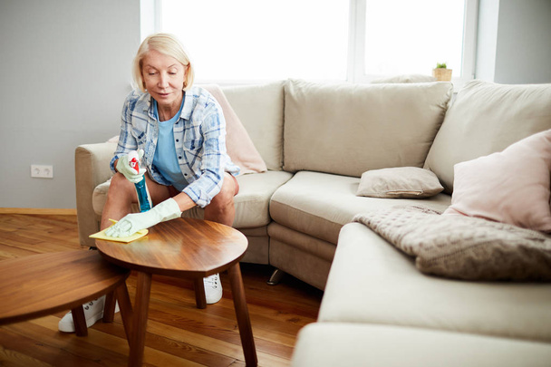 Content attractive mature woman in casual clothing sitting on sofa and using napkin and cleaning supply while removing dust from coffee table - Foto, imagen