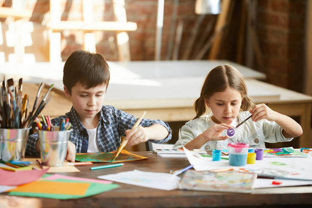 Portret van twee kinderen jongen en meisje schilderen samen zittend aan tafel thuis, kopieer ruimte - Foto, afbeelding