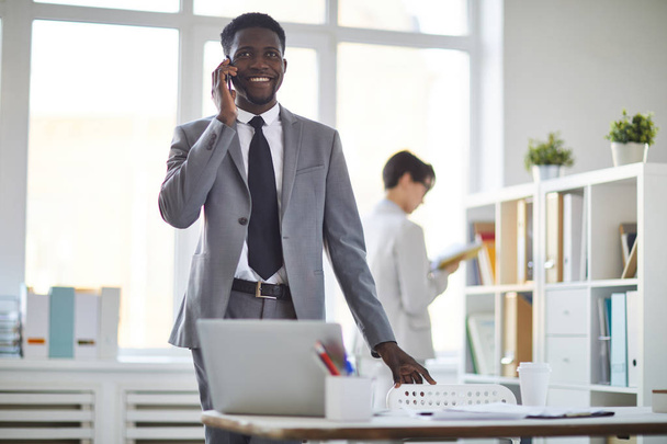 Felice giovane uomo d'affari elegante in giacca e cravatta parlare su smartphone mentre in piedi sul posto di lavoro in ufficio
 - Foto, immagini