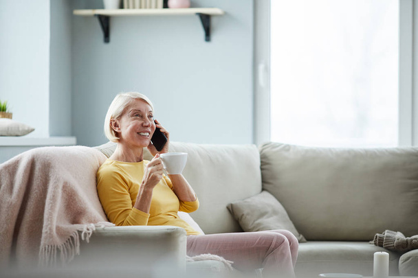 Pozytywna atrakcyjna dojrzała dama z krótkimi włosami siedzi na kanapie i rozmawia na telefon komórkowy podczas picia kawy w domu - Zdjęcie, obraz