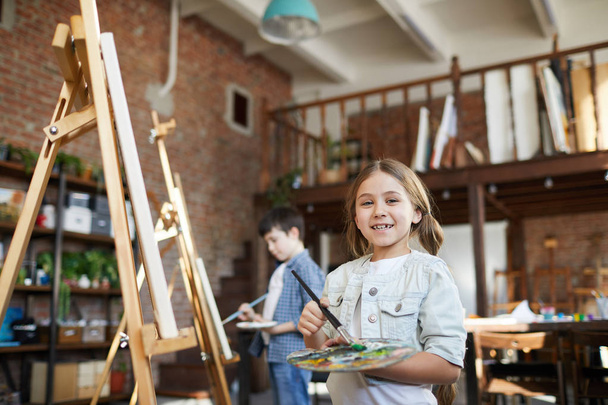 Μέση επάνω πορτρέτο του χαριτωμένο μικρό κορίτσι ζωγραφική εικόνα σε καβαλέτο στην κλάση τέχνης και κοιτάζοντας την κάμερα, αντίγραφο χώρου - Φωτογραφία, εικόνα
