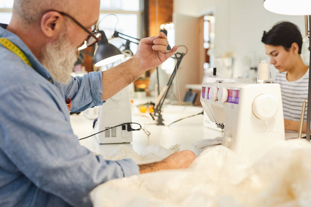 Senior man met naald en draad werken over nieuwe item van de mode-collectie in zijn atelier of Studio - Foto, afbeelding