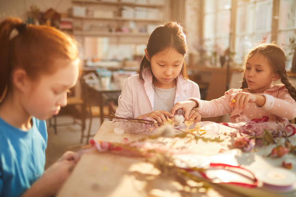 Grupo multiétnico de niñas pequeñas que crean iras de primavera y decoraciones hechas de flores en clase de arte, escena iluminada por la luz del sol serena
 - Foto, Imagen