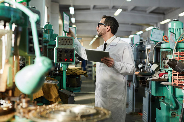 Zijaanzicht portret van volwassen kwaliteit inspecteur dragen lab jas controleren industriële machine units in Factory Workshop, kopieerruimte - Foto, afbeelding