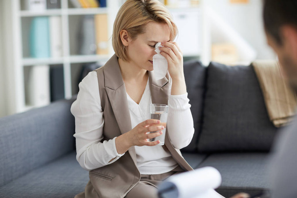 Surullinen ja masentunut nuori nainen pyyhkii kyyneleensä ja ottaa lasillisen vettä istuessaan sohvalla psykologi toimistossa
 - Valokuva, kuva