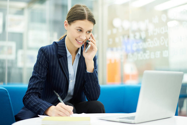 Portrait of smiling young businesswoman speaking by phone in office during internship, copy space - Фото, зображення