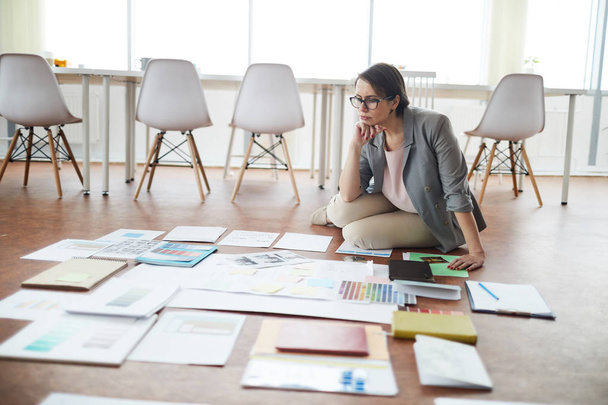 Ganzkörperporträt einer nachdenklichen Geschäftsfrau, die im Büro auf dem Boden sitzt, während sie ein Projekt plant, Kopierraum - Foto, Bild
