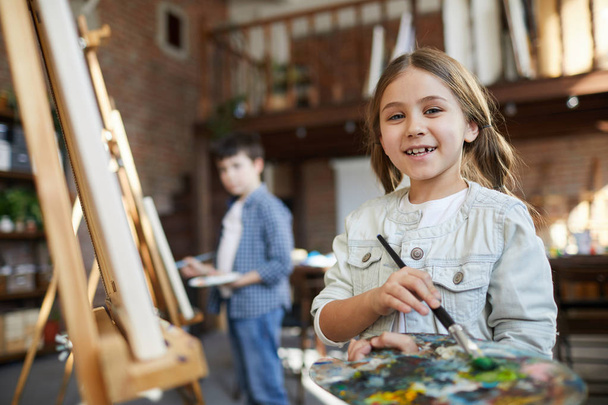 Waist up portrait of cute little girl painting picture on easel in art class and holding palette, copy space - 写真・画像