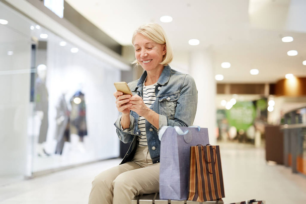 Feliz mujer madura emocionada con el pelo rubio sentado en el banco con bolsas de compras y el uso de teléfono inteligente al hacer post sobre las compras en las redes sociales
 - Foto, imagen