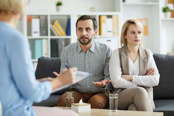 Offended woman with crossed arms looking aside while her husband trying to explaining their problem to counselor - Photo, Image